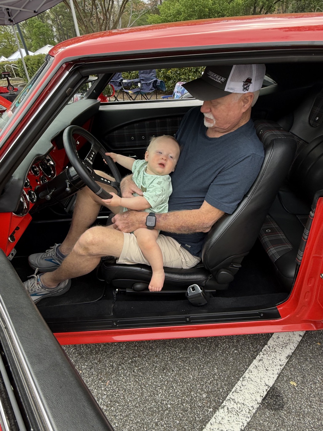 Morgan family member in a Morgan Muscle Cars cap with his grandchild in a red muscle car at an Alabama classic car show.